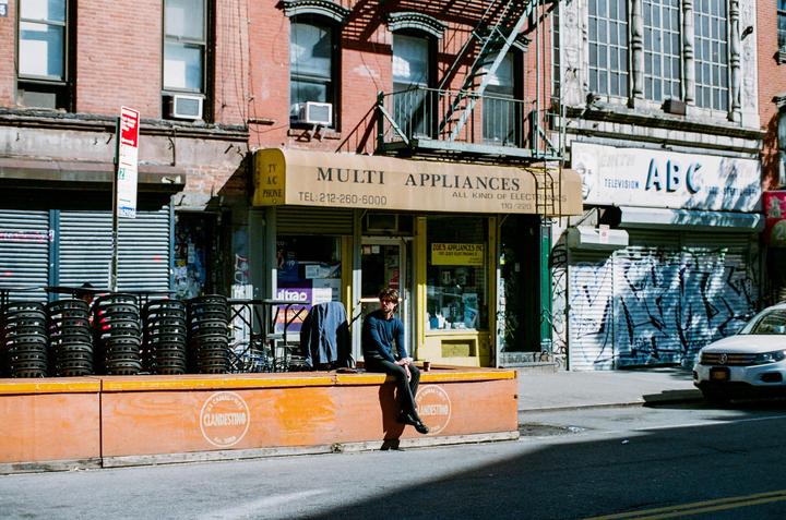 A man sits on a low orange wall in front of a store with a yellow awning that says "Multi Appliances" on a sunny street.