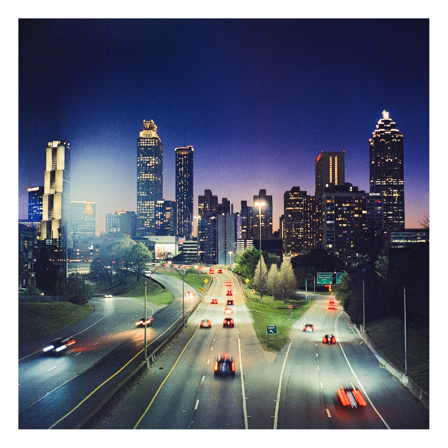 A highway with blurred car lights leads into a city skyline at dusk.