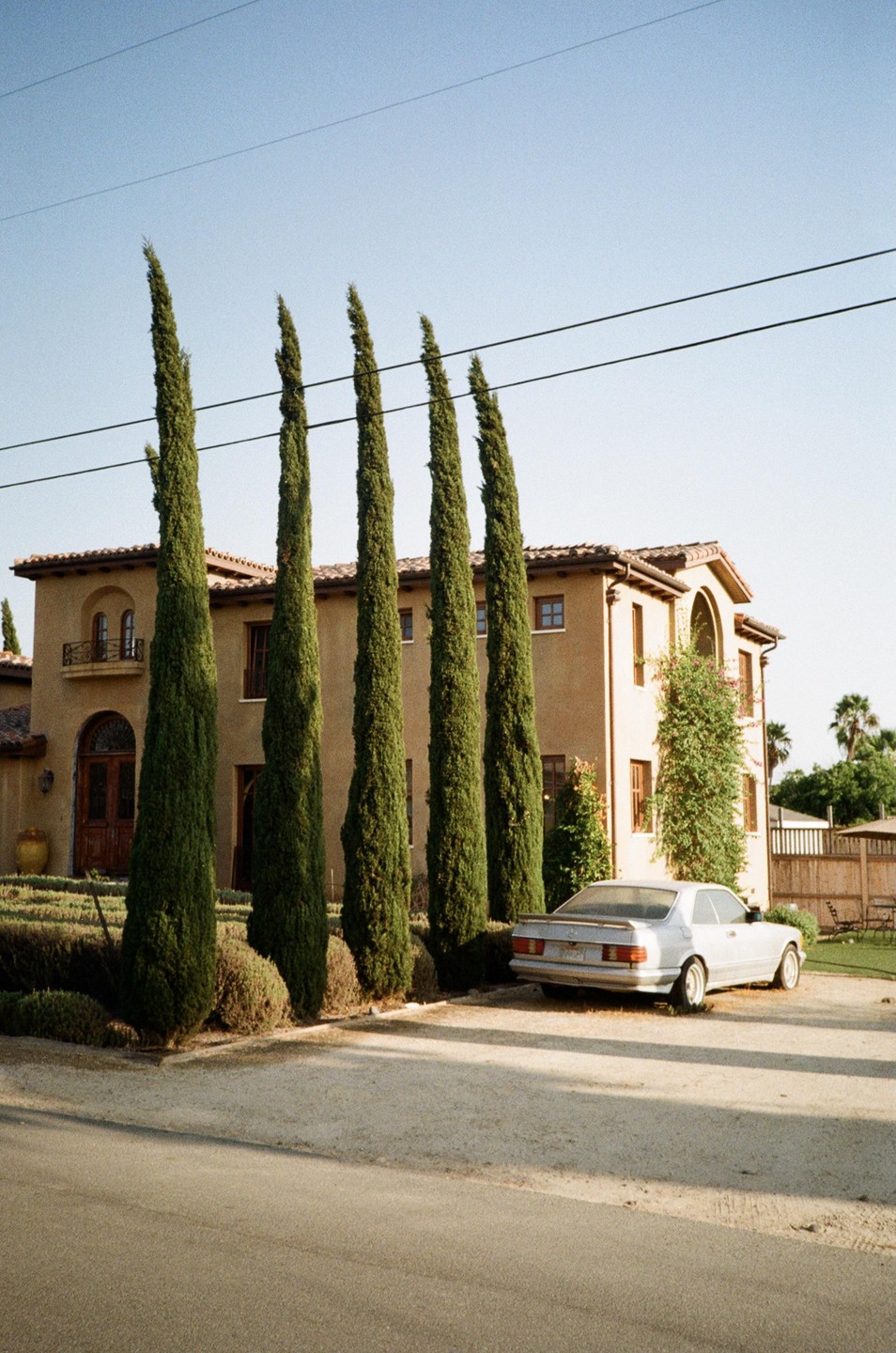 A silver car is parked in front of a large house with tall, narrow trees lining the driveway.