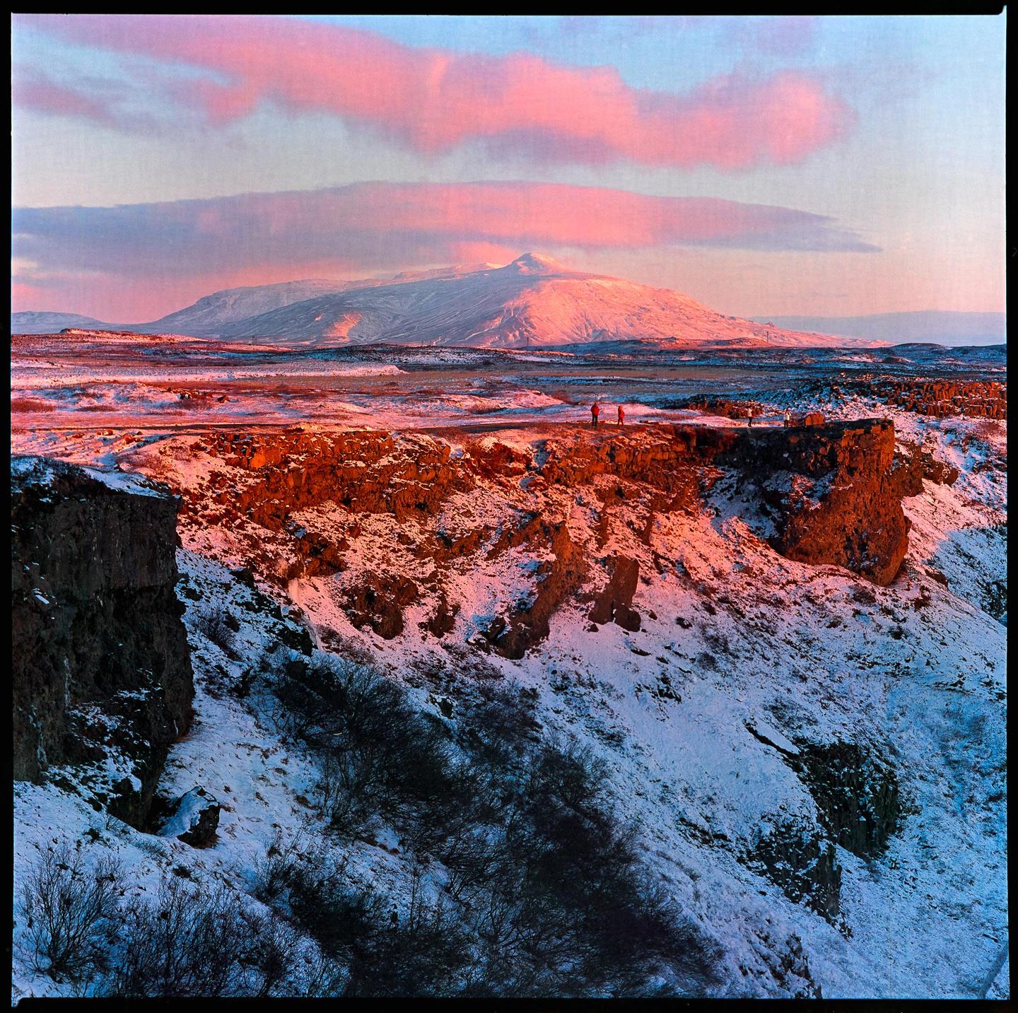 Two people stand on a snowy cliff overlooking a vast, mountainous landscape under a sky with pink clouds.