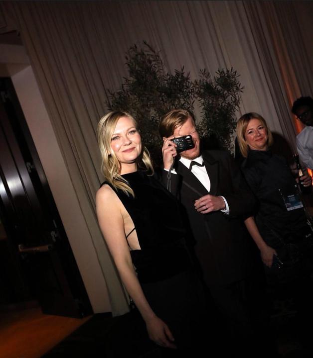 A woman in a black dress smiles at the camera while a man next to her holds a camera to his face, and another woman stands to their right.