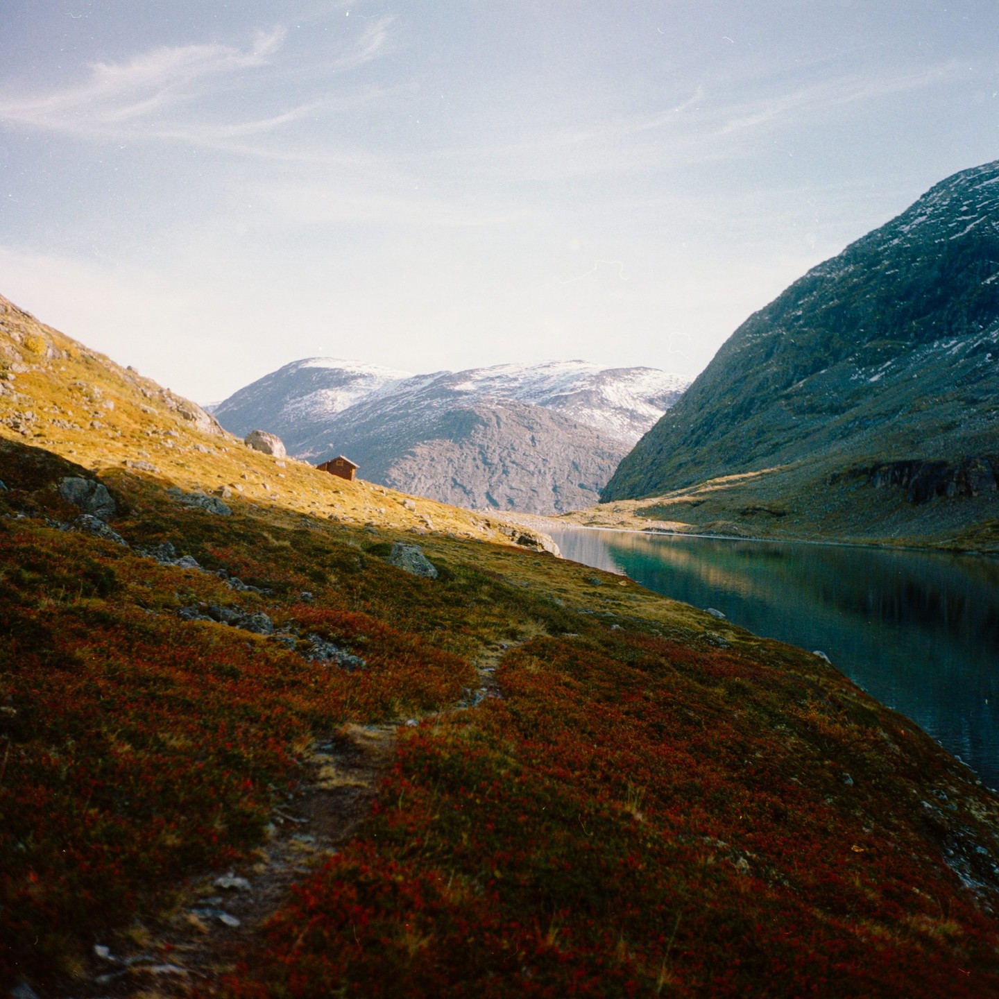 A small cabin sits on a hill overlooking a lake surrounded by mountains, with a path leading towards it.