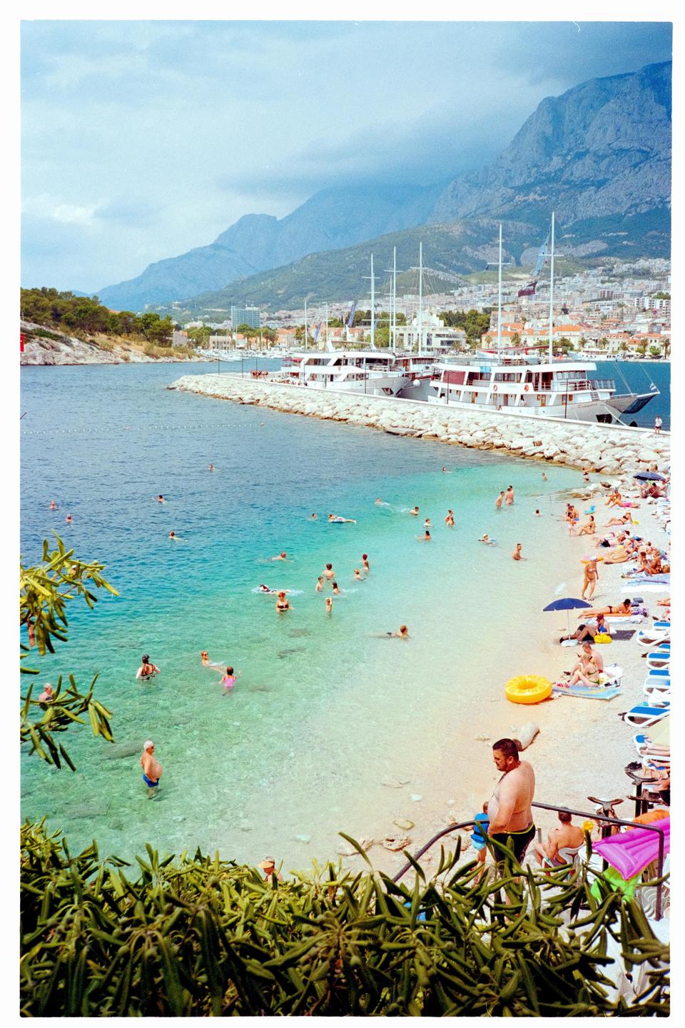 A crowded beach with people swimming in clear blue water and sunbathing on the sand, with boats docked at a pier and mountains in the background.