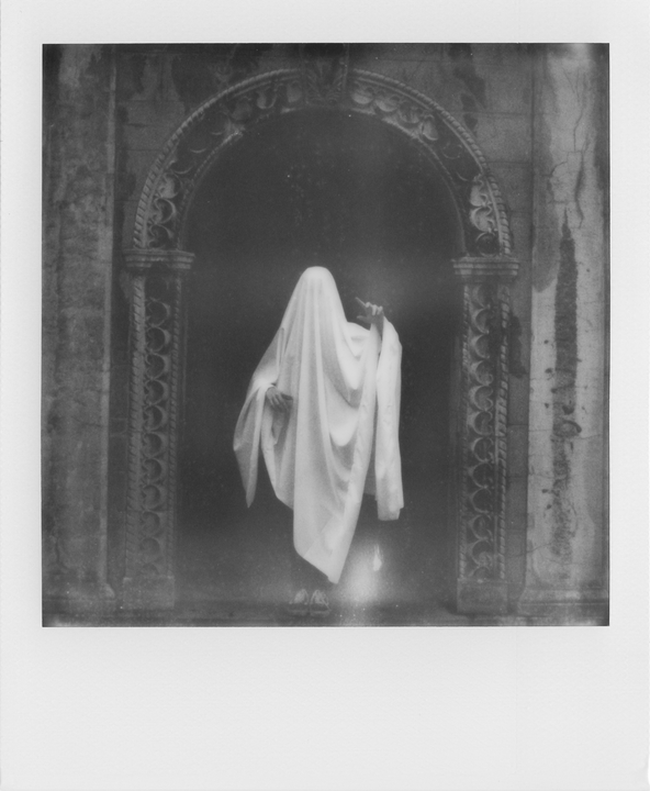 A person covered in a white sheet stands in front of an ornate archway.