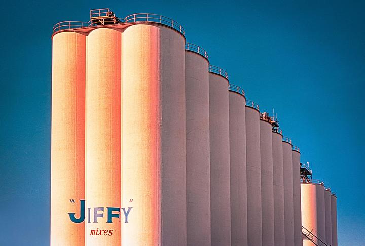 A row of tall, cylindrical silos with "Jiffy mixes" written on the first one stands against a clear blue sky.