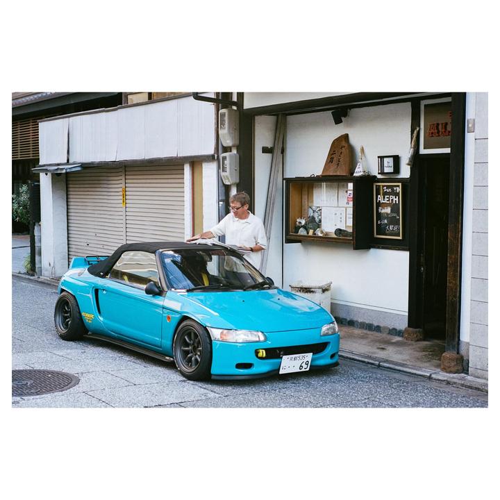 A man in a white shirt stands next to a turquoise convertible car parked on a narrow street.