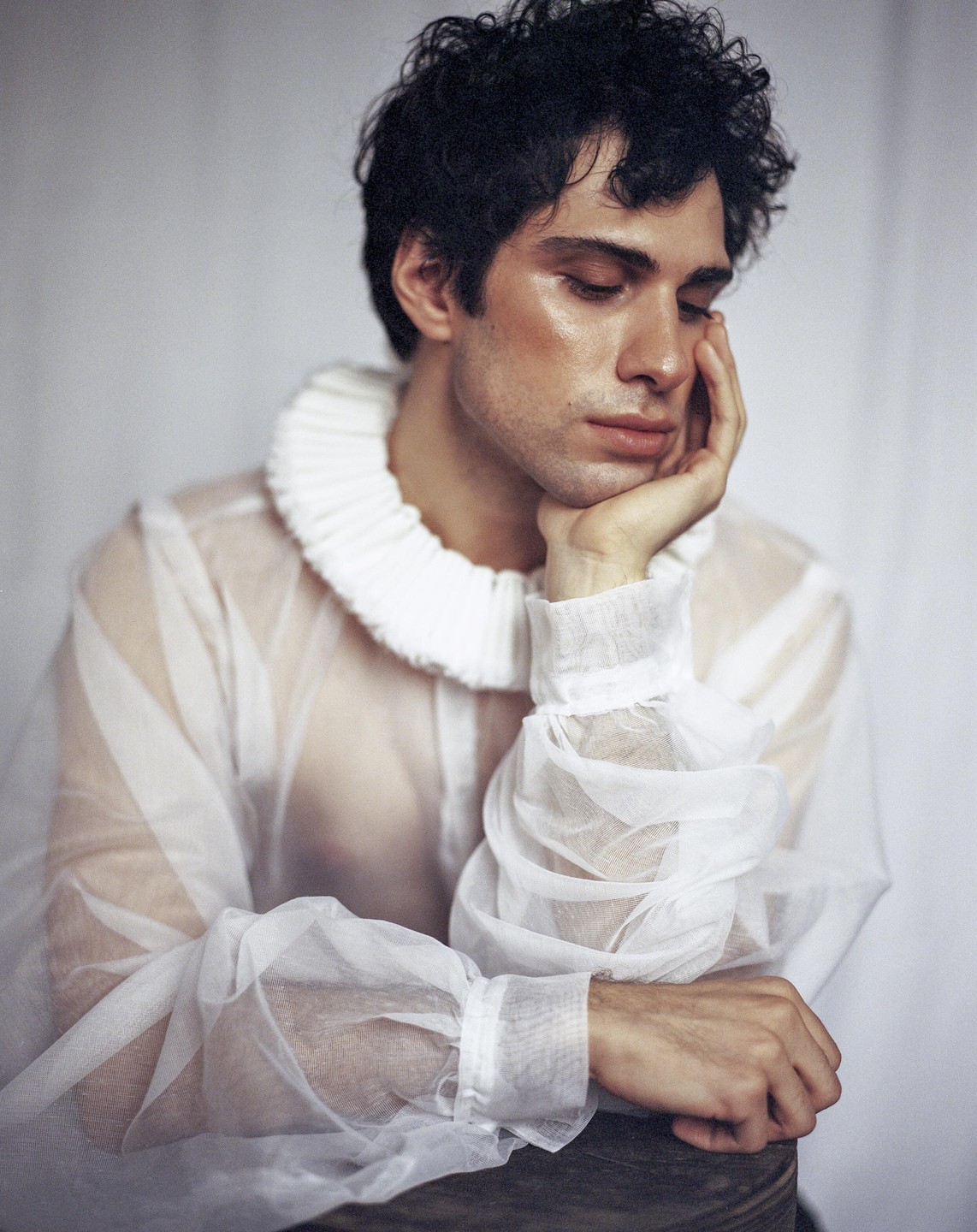 A man with dark curly hair wears a sheer white ruffled shirt and rests his head on his hand.