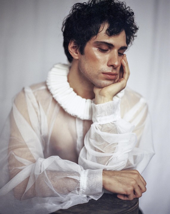 A man with dark curly hair wears a sheer white ruffled shirt and rests his head on his hand.