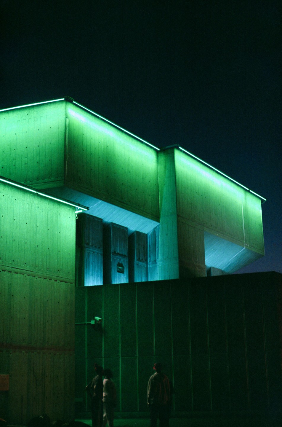 Three people stand in front of a building illuminated with green and blue neon light at night.