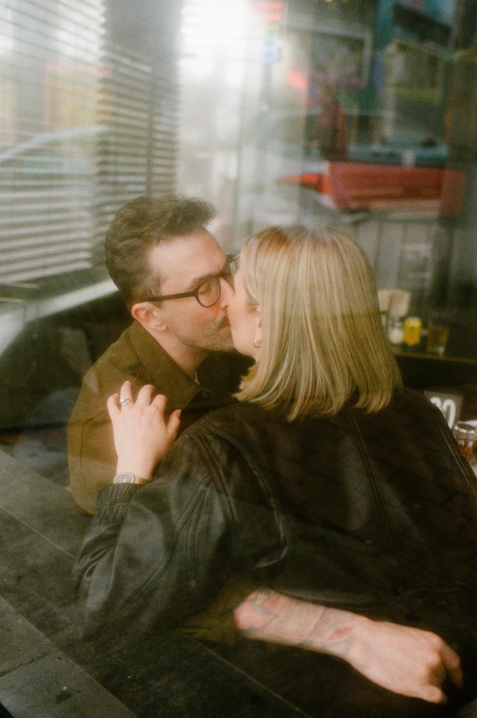 A couple kisses through a window, with reflections of a city street visible in the glass.