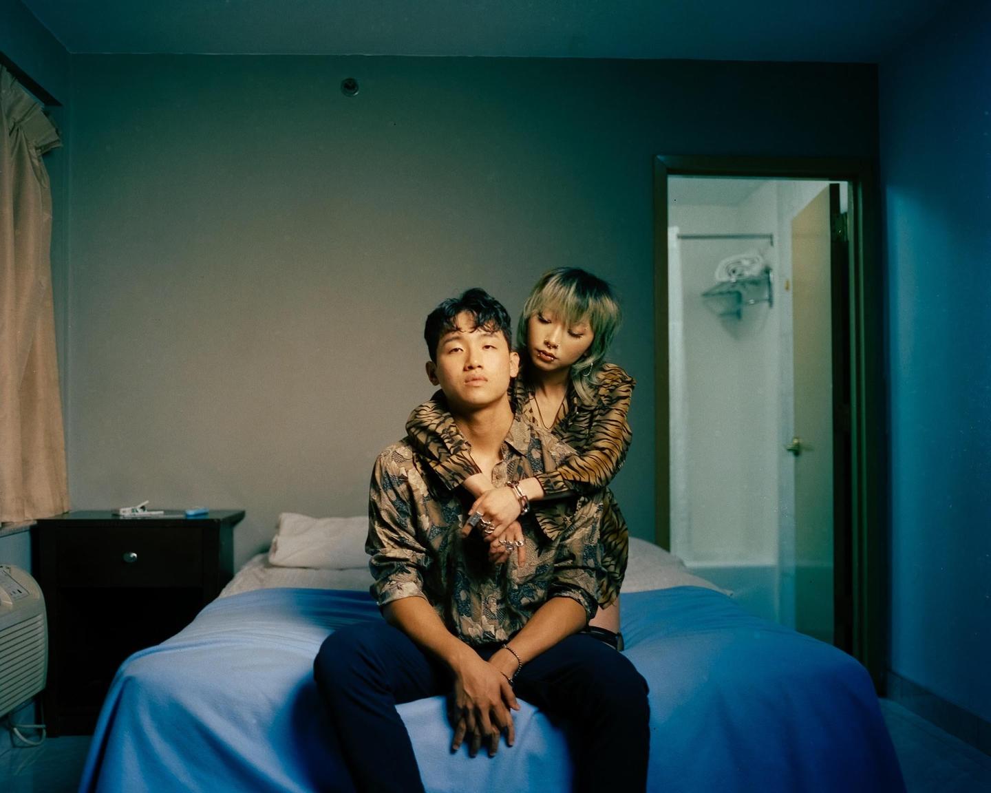 A couple sits on a bed in a dimly lit room, with the woman embracing the man from behind.