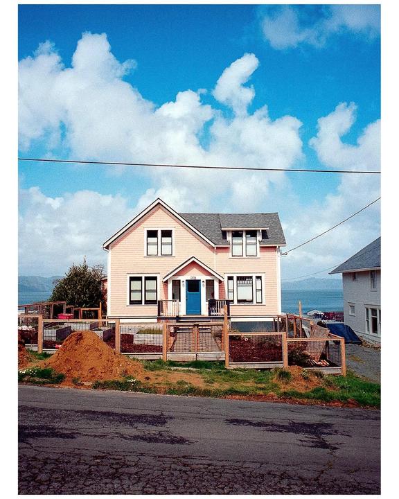 A pink house with a blue door sits on a hill overlooking a body of water under a bright blue sky with white clouds.