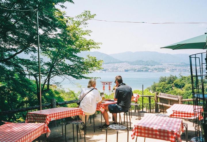 A couple sits at a cafe table with a view of the water and a torii gate.