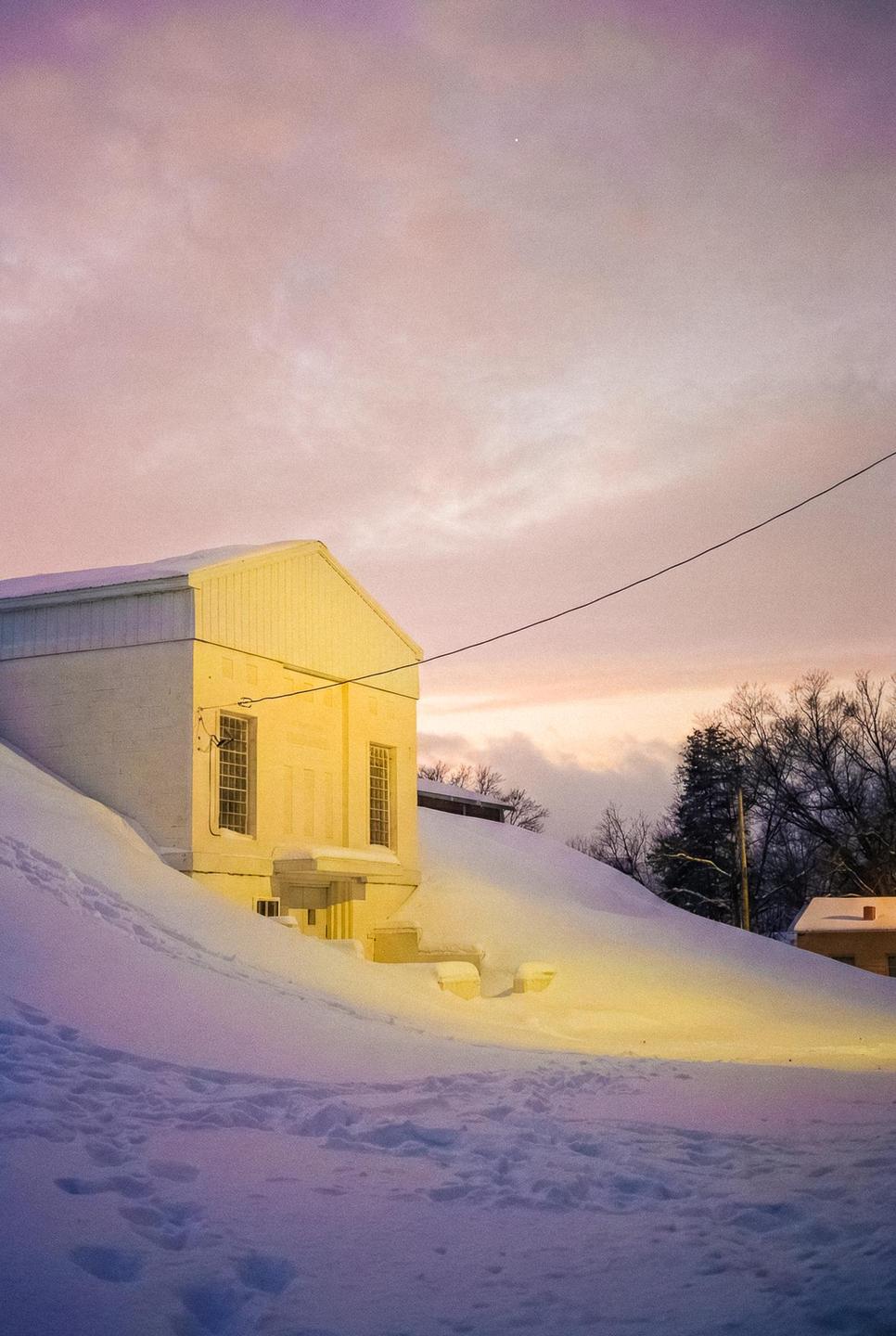A building is surrounded by deep snow under a colorful sky at dusk.