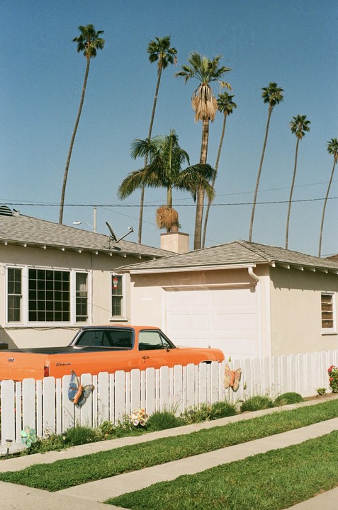 An orange car is parked behind a white picket fence in front of a house with palm trees under a clear sky.