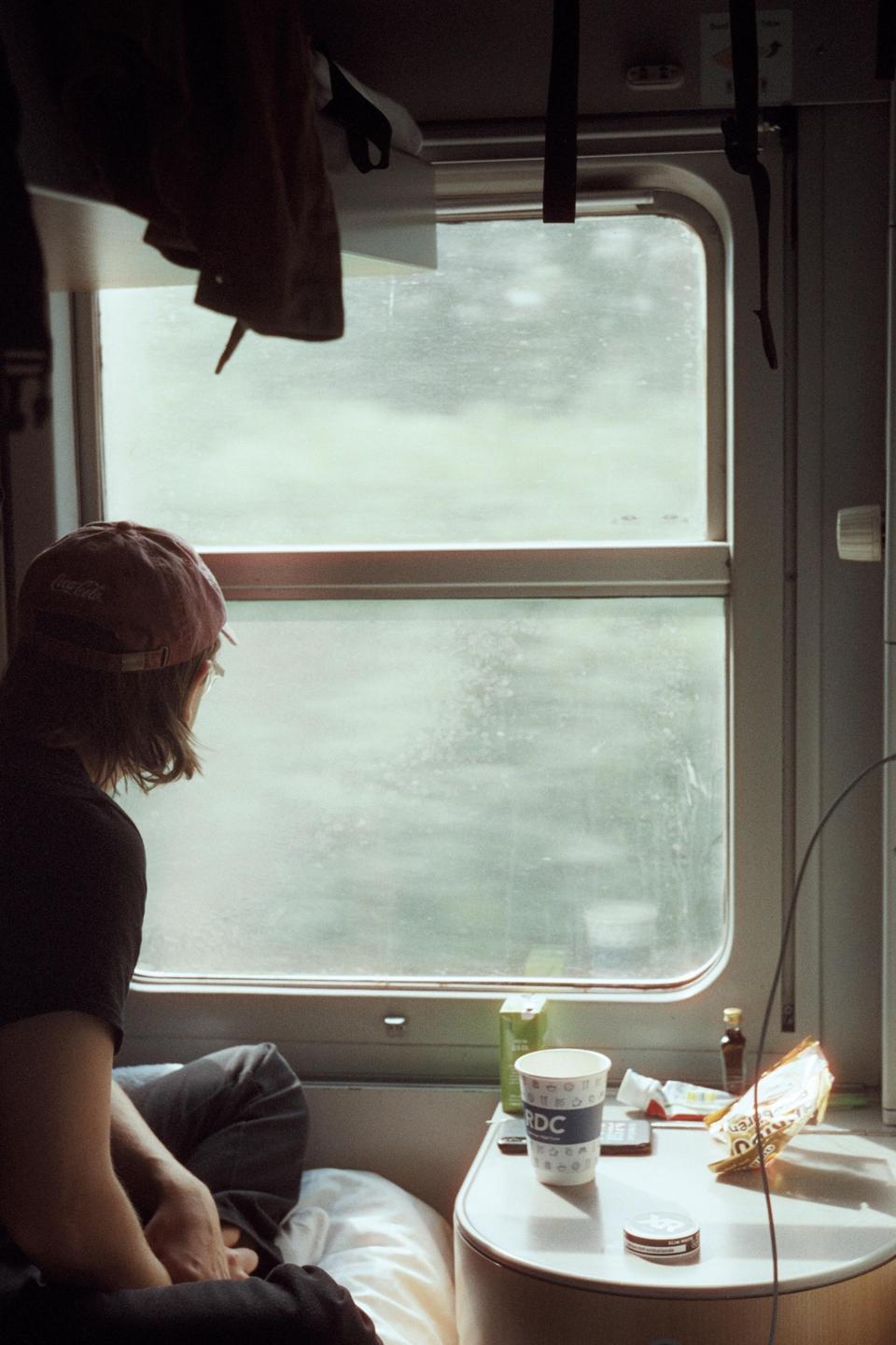 A person wearing a baseball cap looks out a train window at a passing landscape.