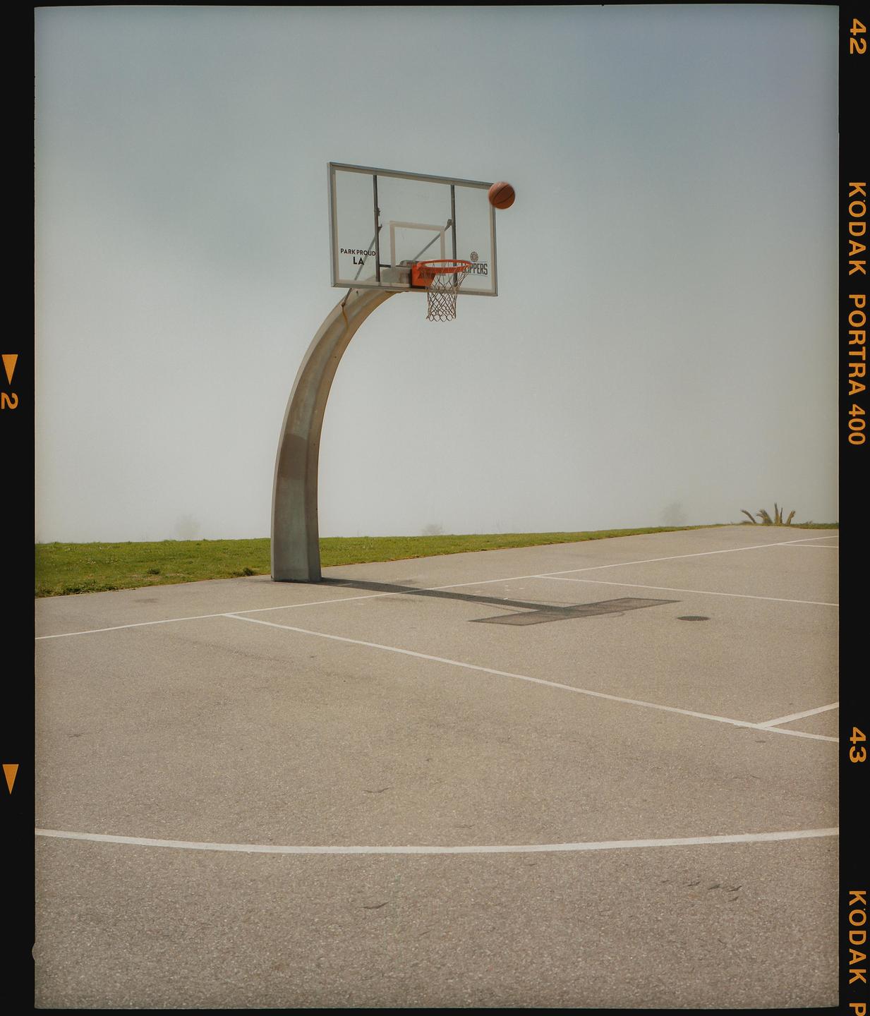 A basketball is in mid-air above a basketball hoop on an outdoor court.