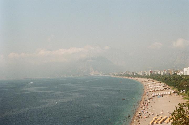 A long beach with many people and umbrellas stretches along a coastline with mountains and buildings in the distance.
