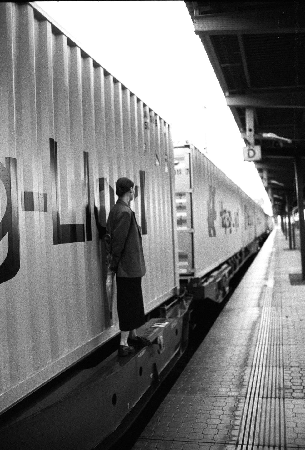 A woman stands on the edge of a train platform next to a long freight train.
