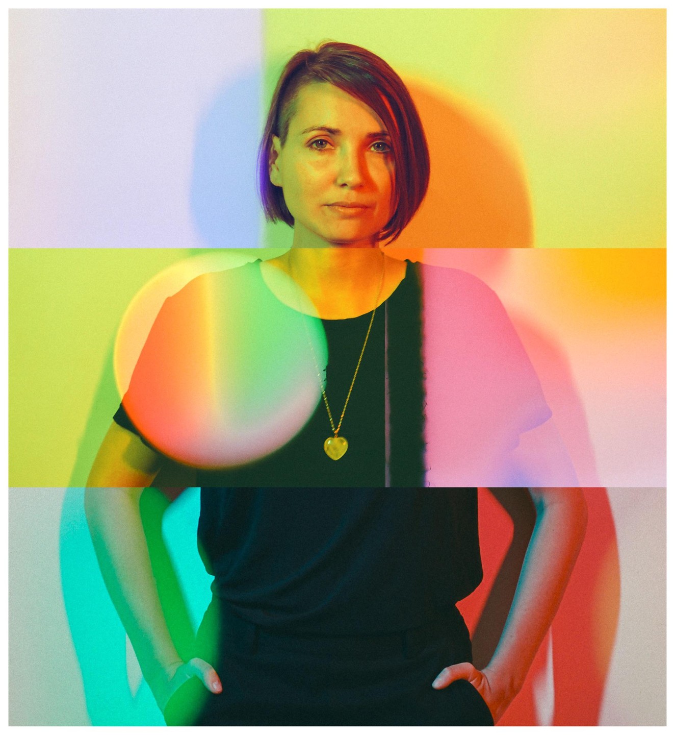 A woman with short hair and a heart pendant stands against a wall with colorful light projections.