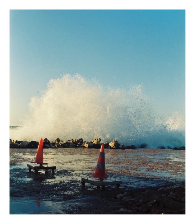 Two orange traffic cones sit on a wet, rocky shore as a large wave crashes behind them.