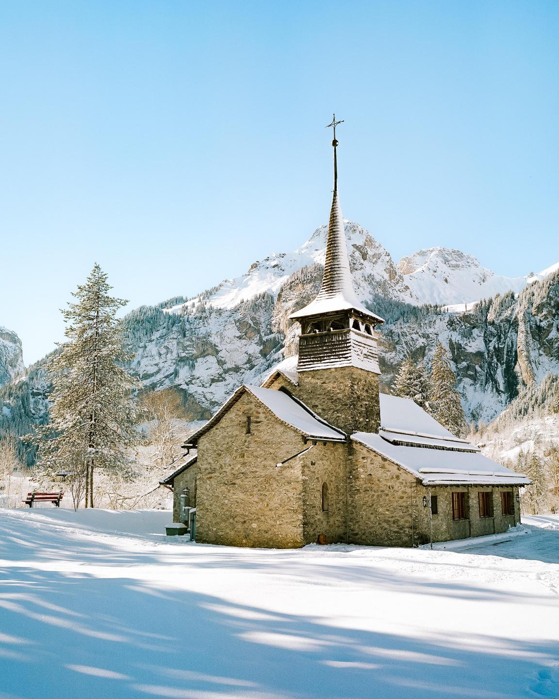 A stone chapel with a tall steeple is covered in snow, set against a backdrop of snow-capped mountains and a clear blue sky.