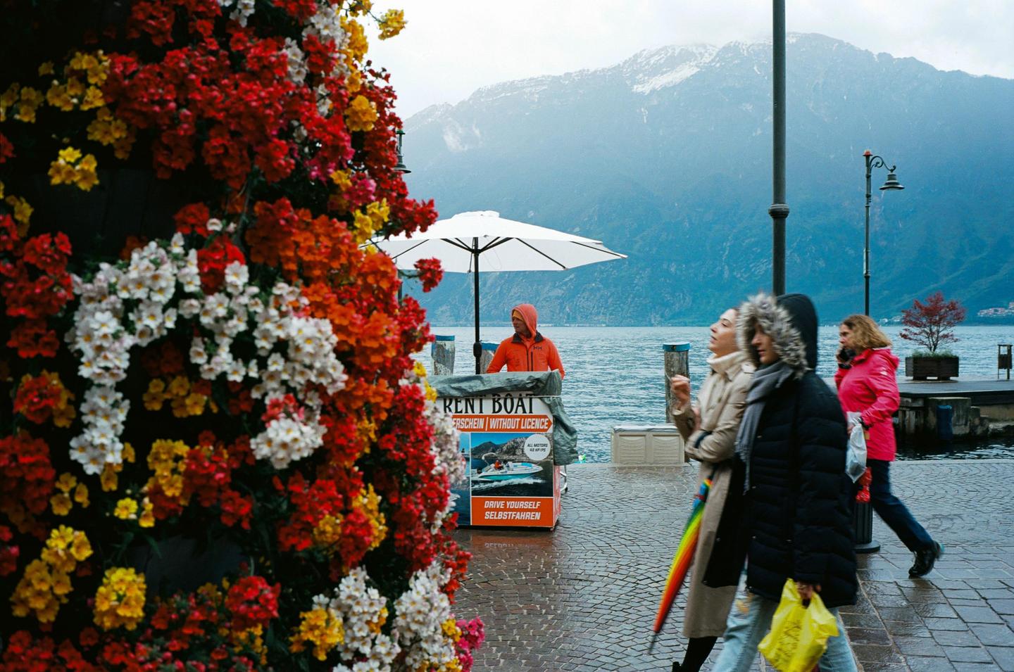 A person in an orange jacket stands by a "rent boat" sign next to a body of water with mountains in the background, while three people walk past a wall of colorful flowers in the foreground.