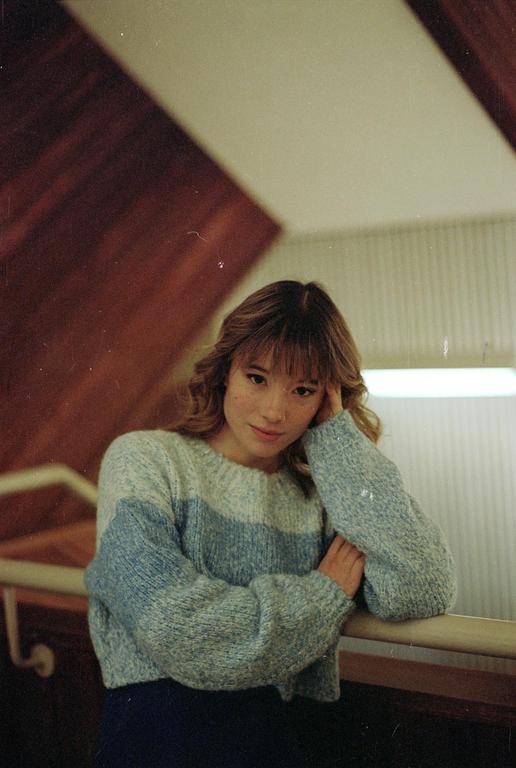 A woman with bangs and a striped sweater leans on a wooden railing.