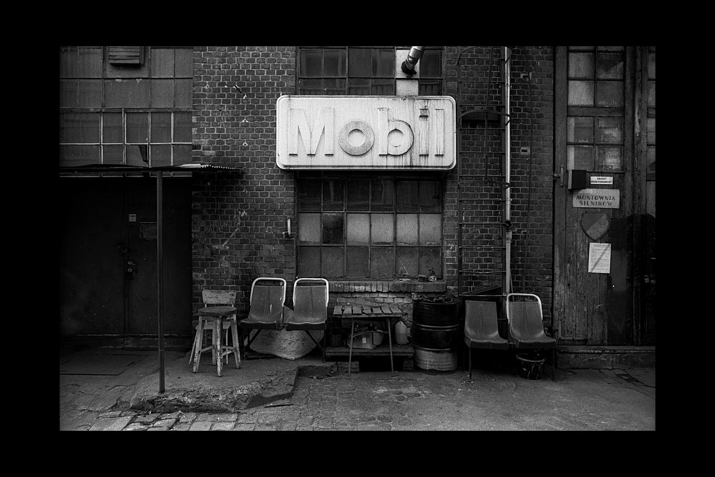 A Mobil sign hangs above a window on a brick building with chairs and barrels outside.