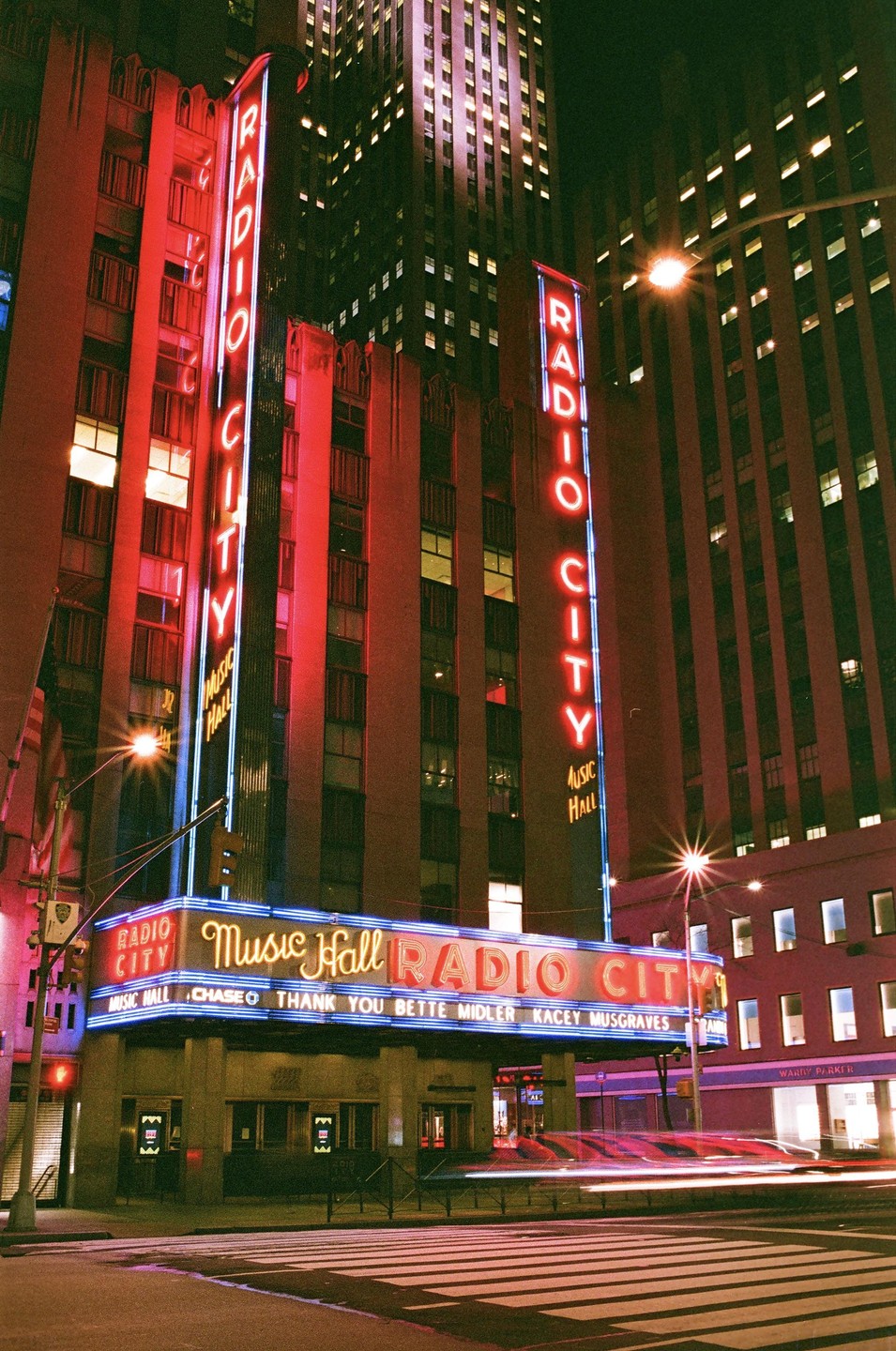 The Radio City Music Hall building is illuminated by neon signs at night, with light trails from vehicles on the street.
