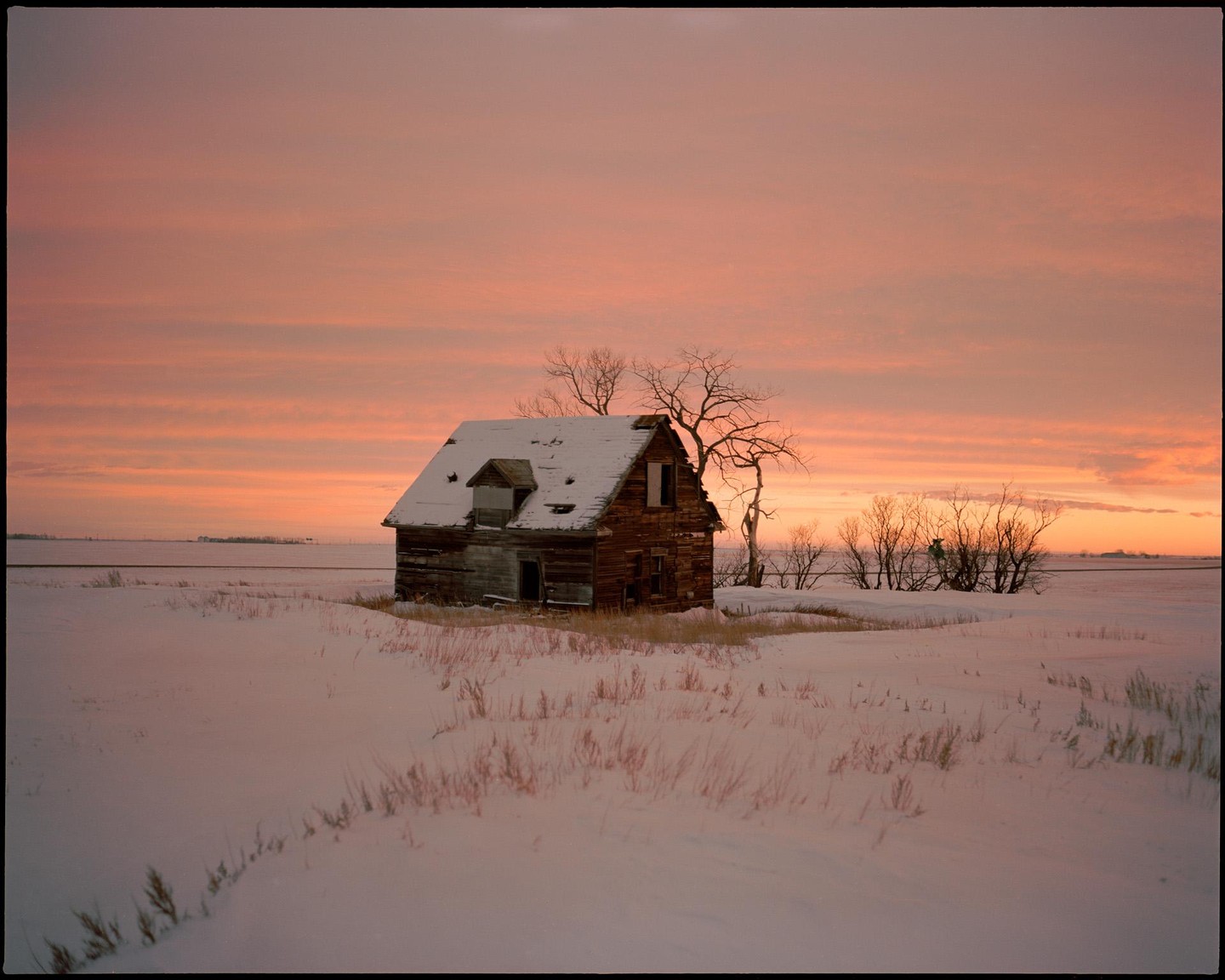 An old wooden house with a snow-covered roof stands in a snowy field under a colorful sunset sky.