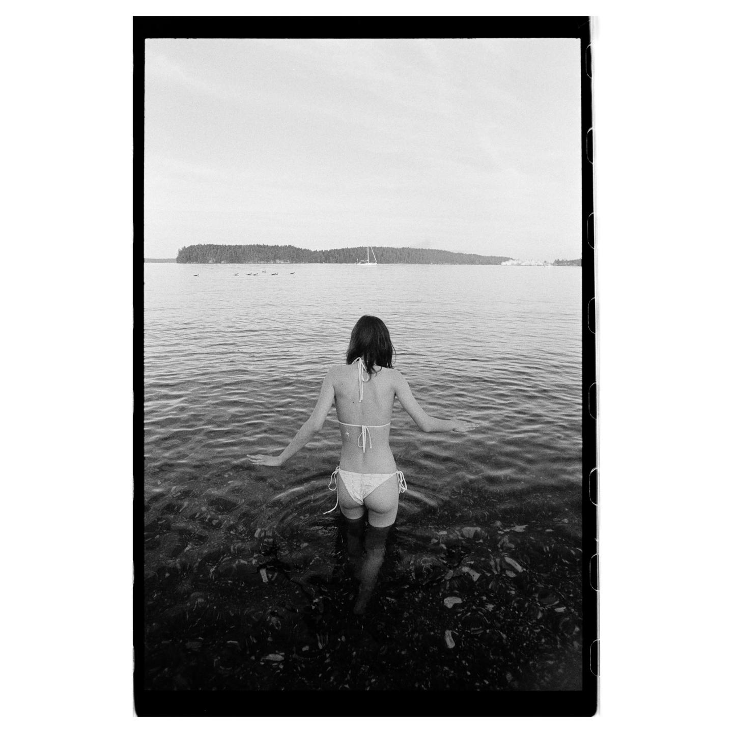 A woman in a bikini stands in shallow water looking out at a body of water with an island in the distance.