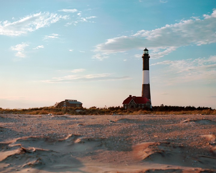 A lighthouse and a small building stand on a sandy beach under a partly cloudy sky.