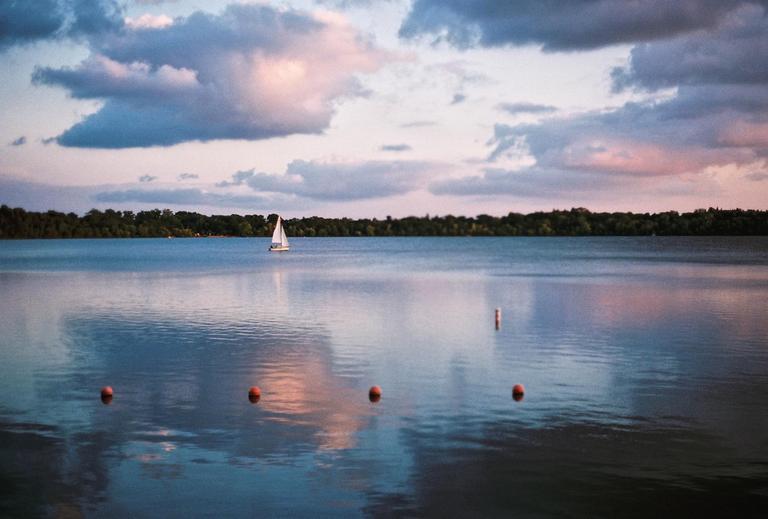 A sailboat floats on a lake with buoys in the foreground and a treeline in the background under a cloudy sky.