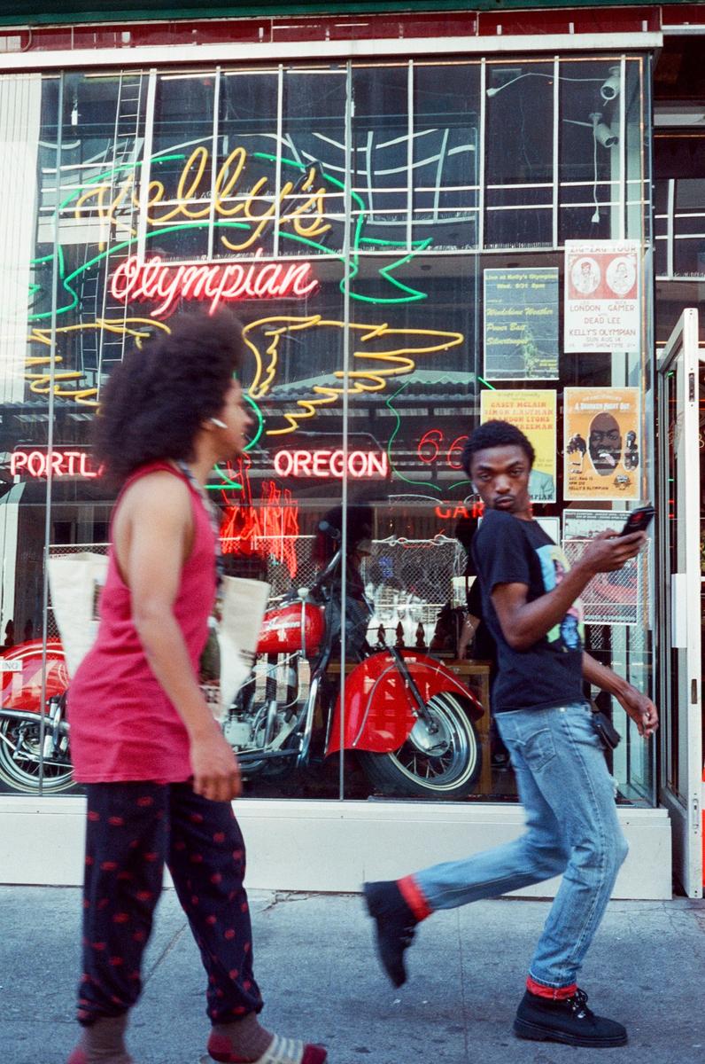Two men walk past a storefront with neon signs and motorcycles displayed in the window.