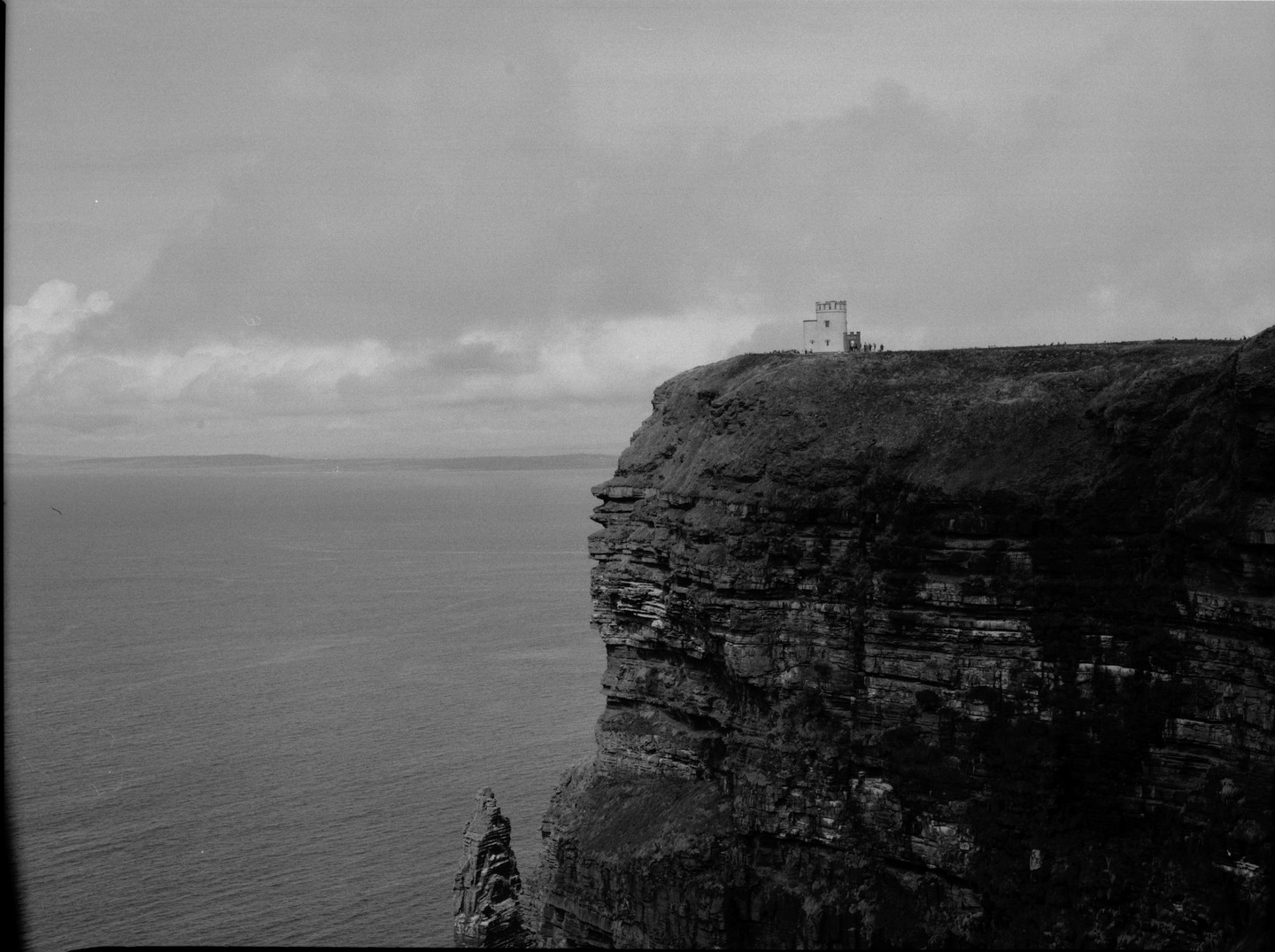 A white tower stands on a grassy cliff overlooking the ocean under a cloudy sky.