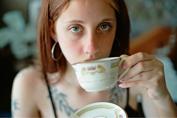 A woman with red hair and a septum piercing drinks from a teacup.