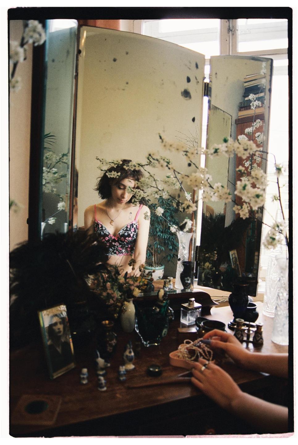 A woman in a floral bra is reflected in a three-panel mirror, surrounded by white blossoms and various trinkets on a wooden dresser.