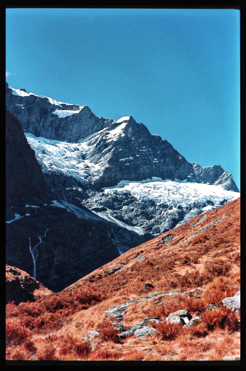 A snow-covered mountain range rises above a valley with red foliage and a small waterfall.