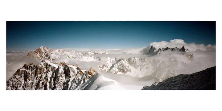 Snow-covered mountain peaks are visible through a layer of clouds under a clear blue sky.