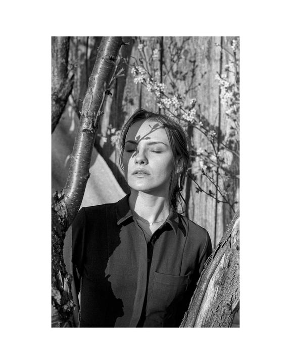 A woman with closed eyes stands between two tree trunks, with shadows of branches on her face and a wooden fence with blossoms in the background.