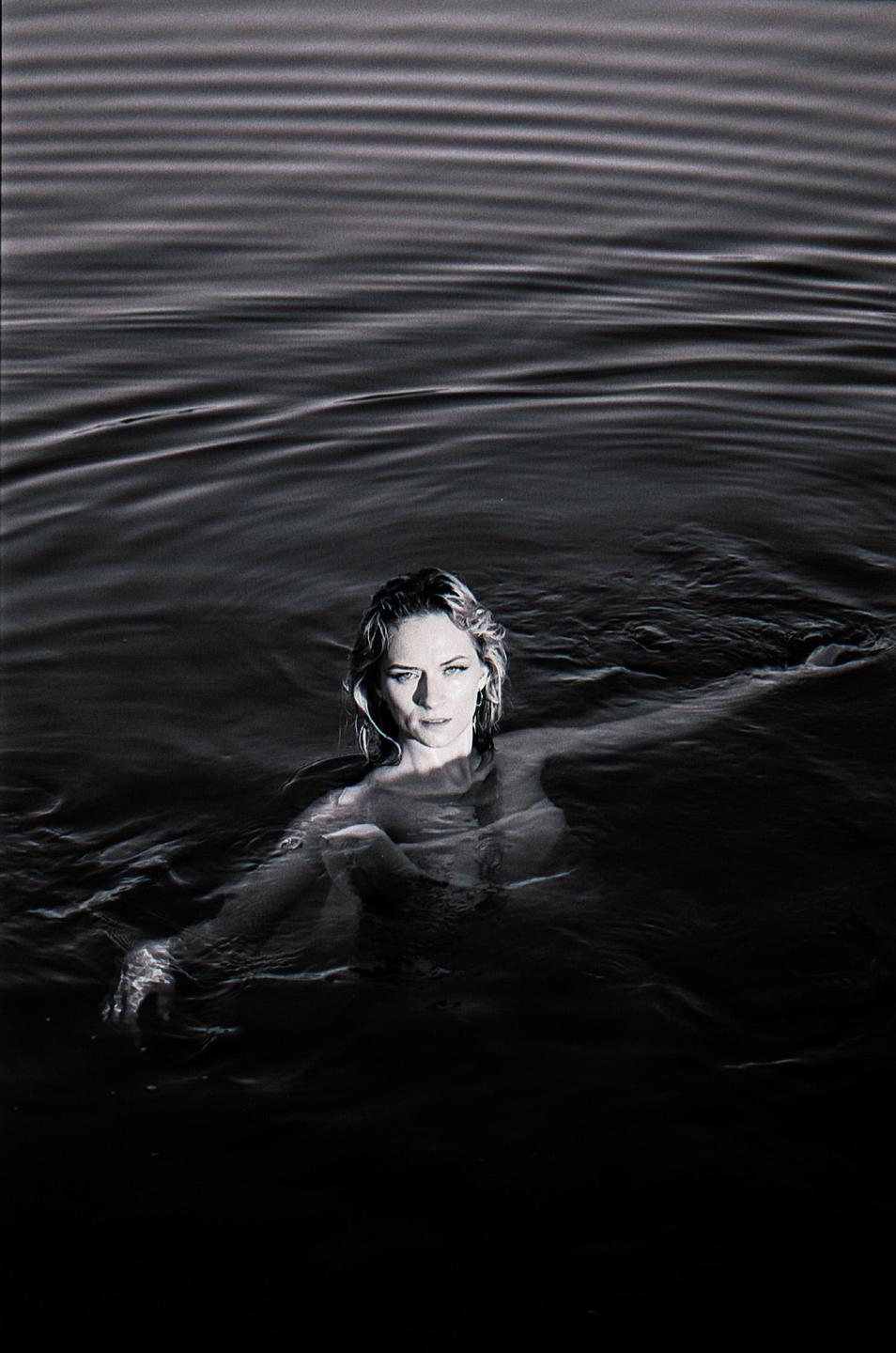 A woman with wet hair is swimming in dark water, looking directly at the viewer.