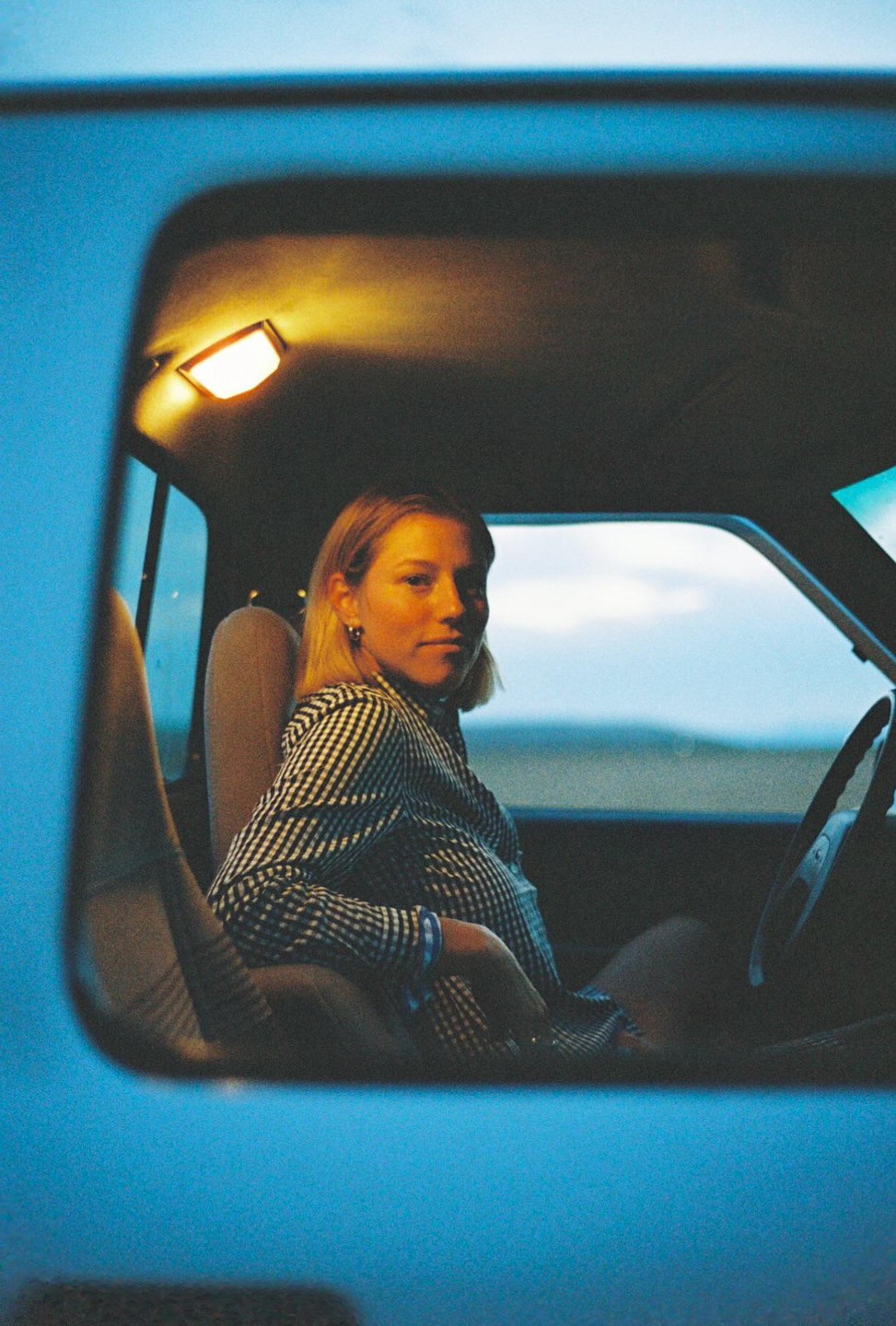 A woman in a checkered shirt sits in a vehicle with an interior light on.
