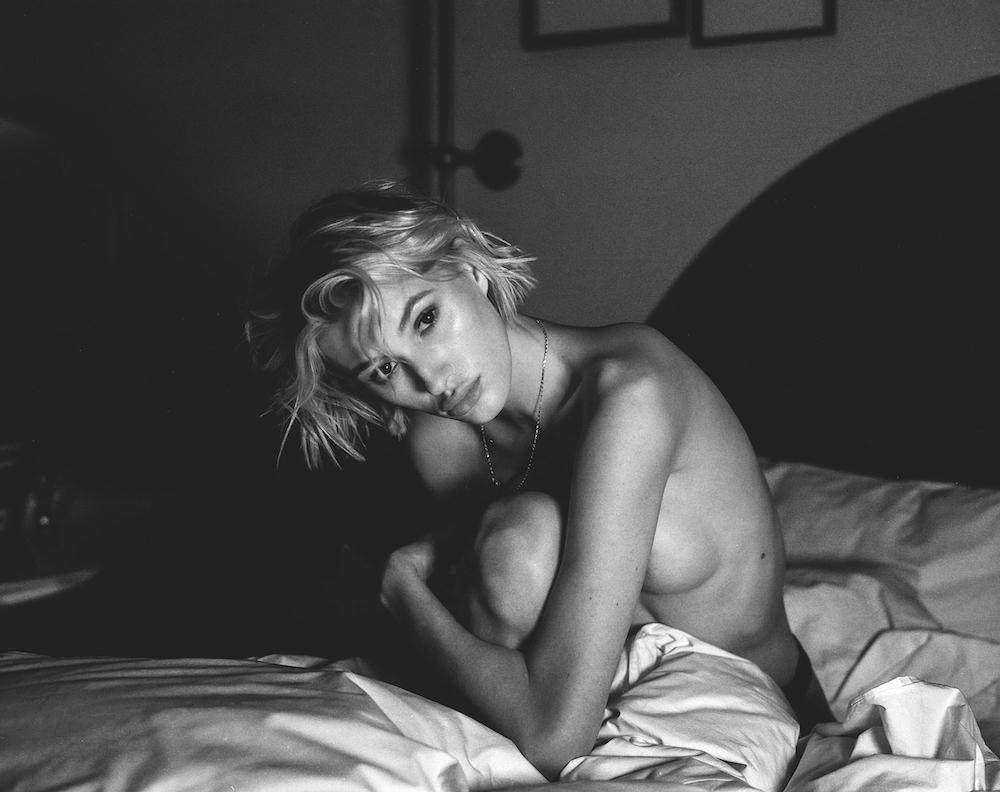 A woman with short hair is sitting on a bed, partially covered by a sheet, and looking at the viewer.
