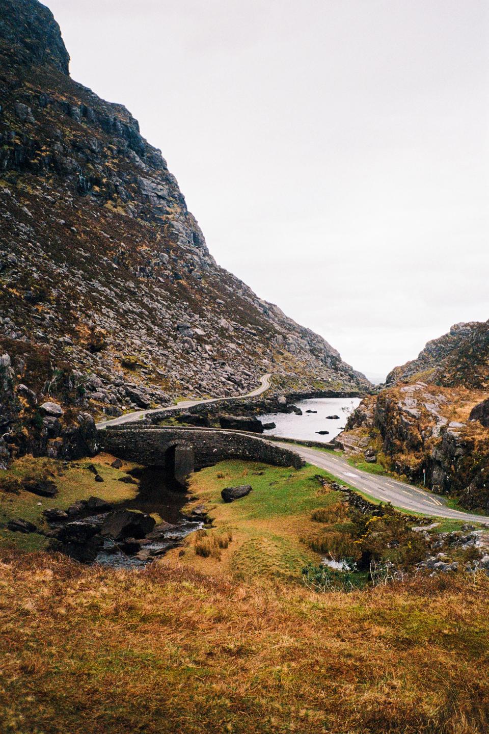 A narrow road winds through a valley with a stone bridge crossing a stream and a body of water.