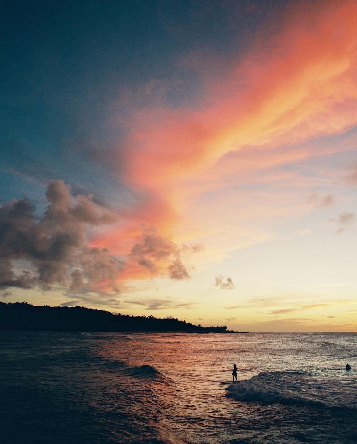 A person stands on a surfboard in the ocean at sunset with a colorful sky and a dark coastline in the background.