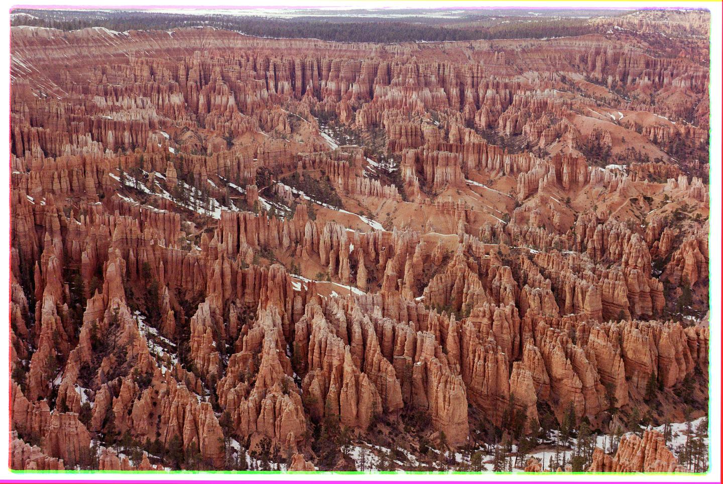 A vast landscape of red rock formations with patches of snow and scattered trees stretches under a distant forest.