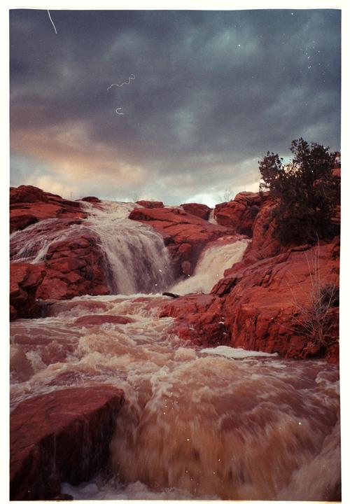 A waterfall cascades over red rock formations under a cloudy sky.