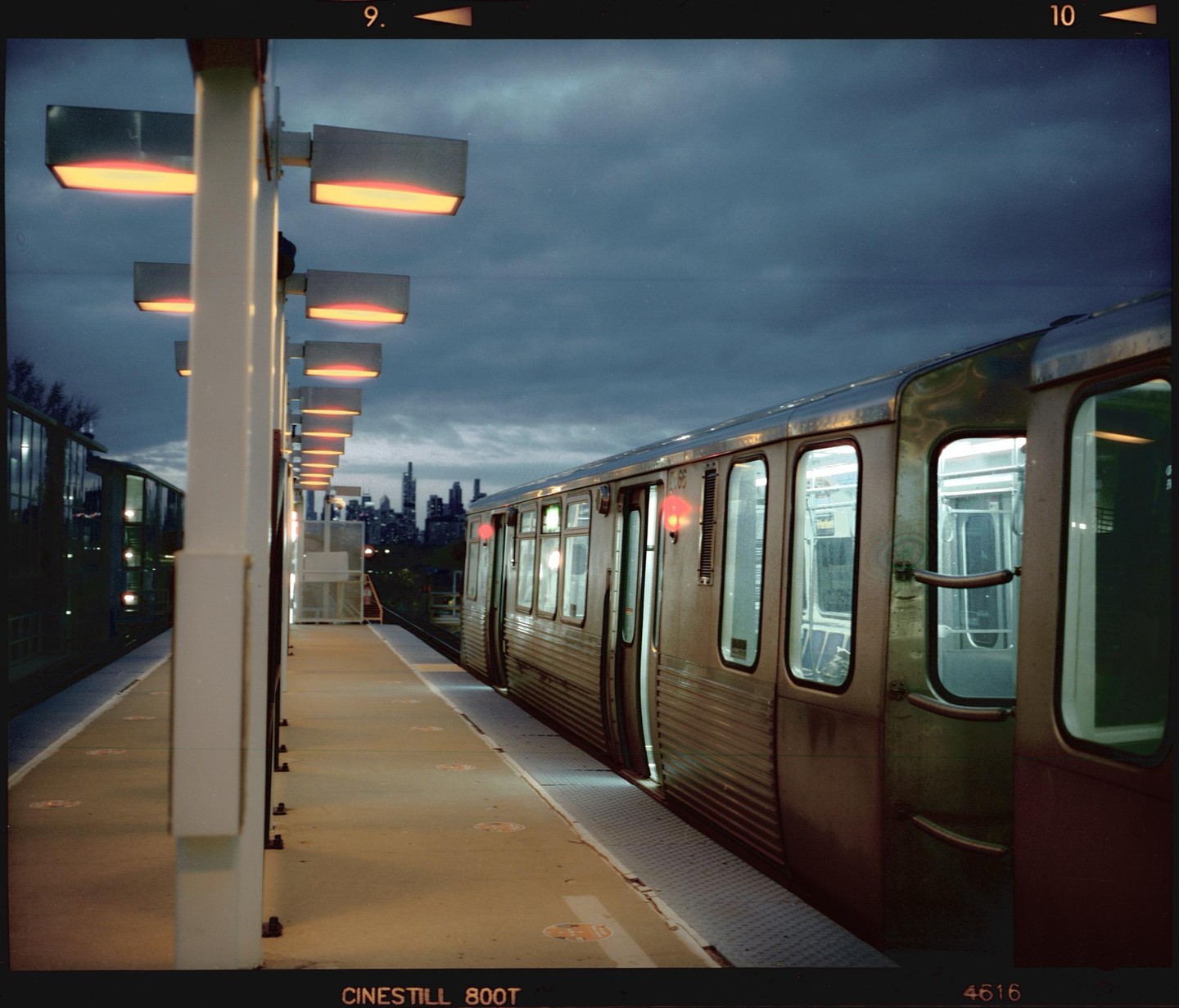 A train waits at a station platform under a cloudy sky, with a city skyline visible in the distance.