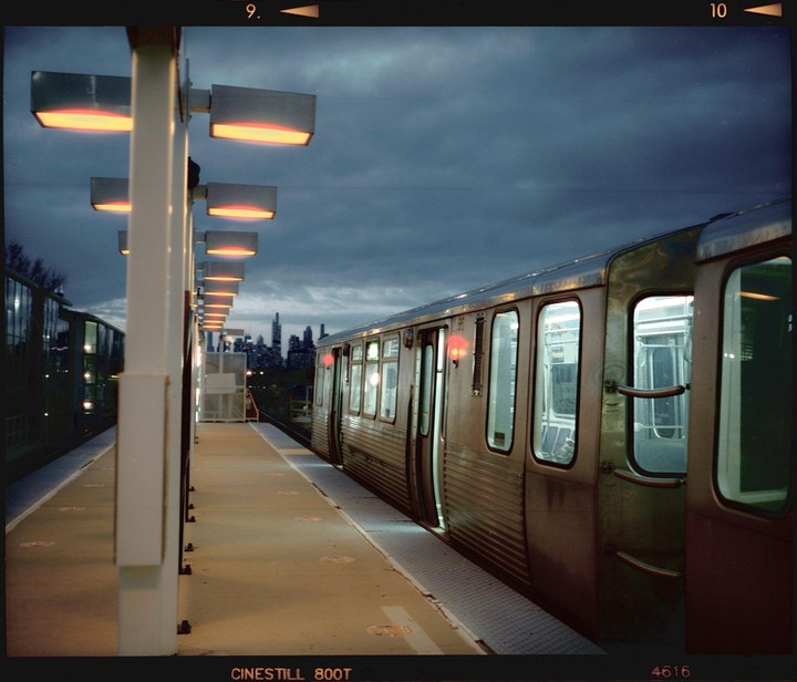 A train waits at a station platform under a cloudy sky, with a city skyline visible in the distance.
